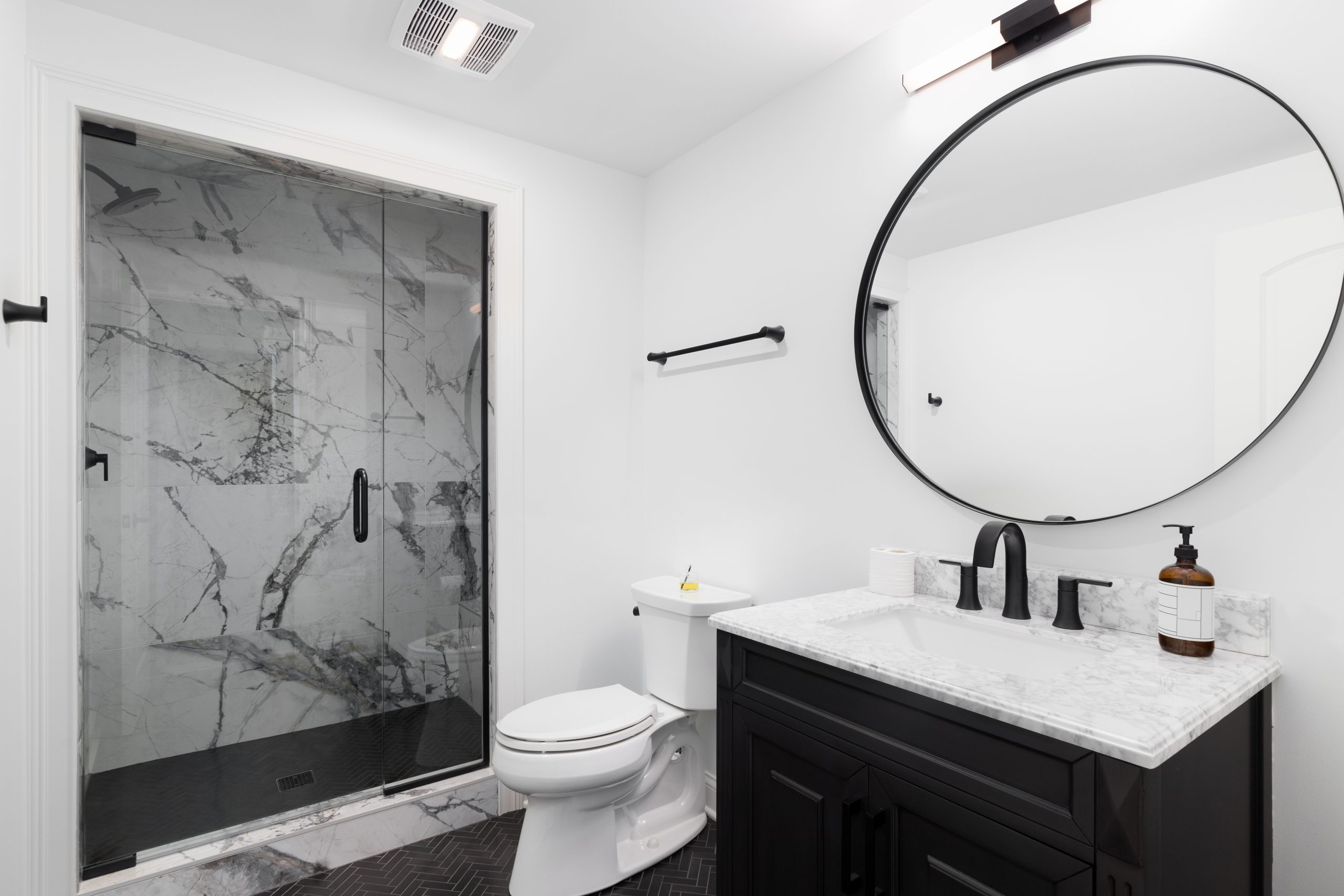 Small bathroom remodel in a North Shore Chicago home featuring a dark wood floating vanity, a large round mirror, herringbone porcelain floor tile, and a frameless glass walk-in shower with marble tiled walls.