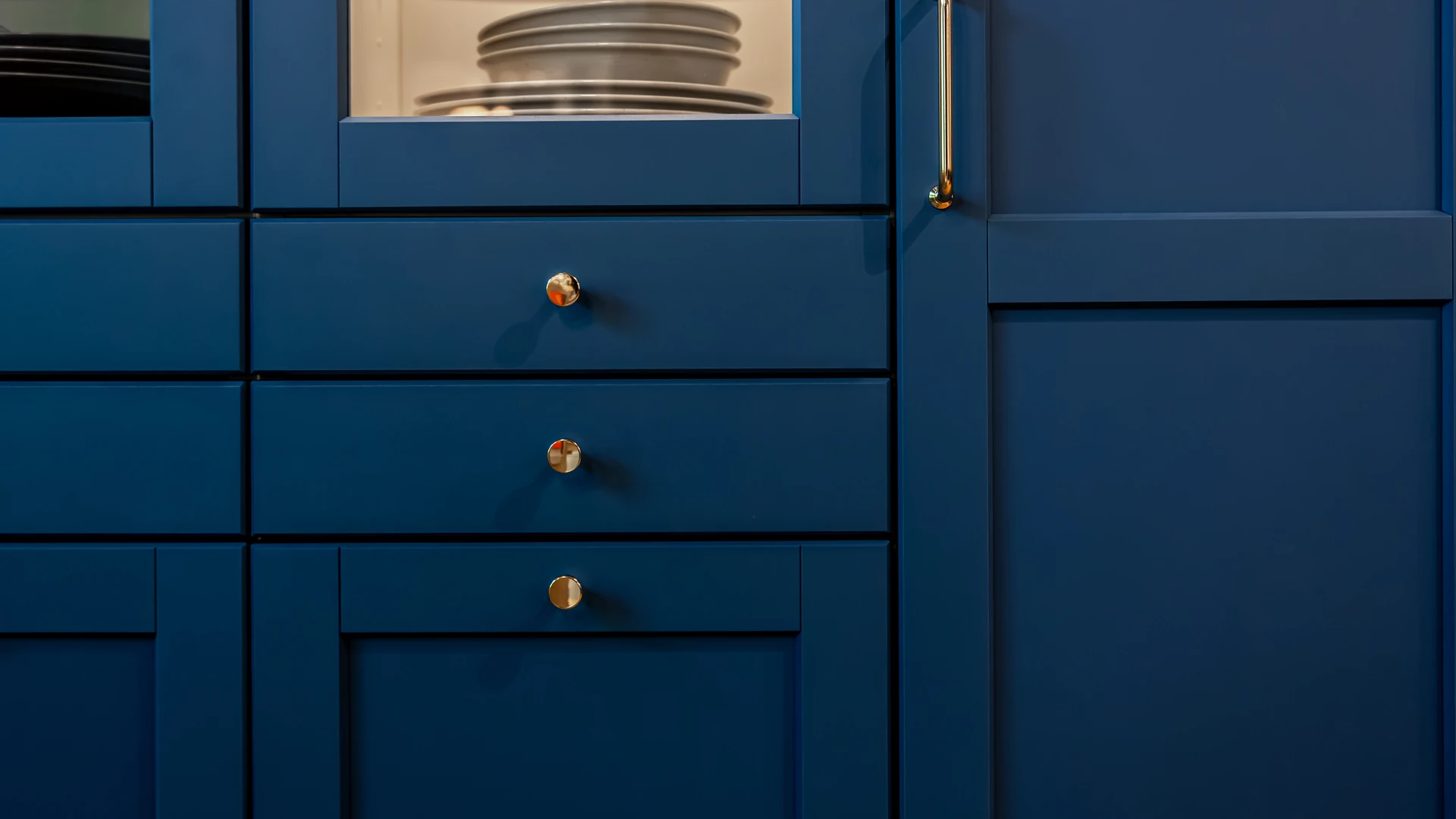 Close-up of navy blue shaker cabinets with brass round knobs and a brass door pull, beside a glass-front cabinet showing stacked plates
