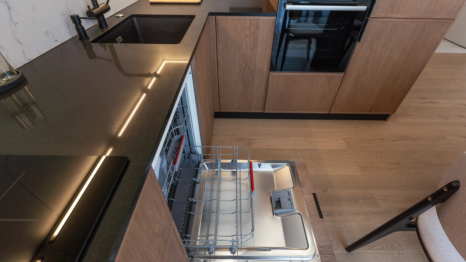 Open built-in dishwasher with its lower rack extended in a dark wood kitchen cabinet run, beside a built-in wall oven and a black granite sink counter