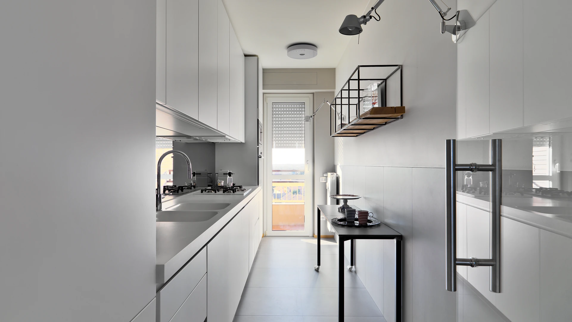 Narrow galley kitchen with white handleless cabinets, a long counter with sink and cooktop, and a slim table opposite