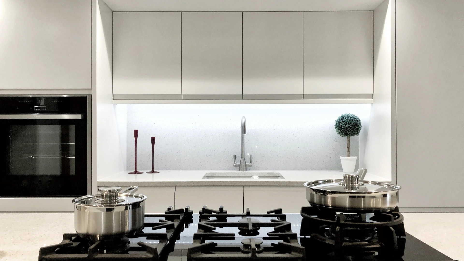 Gas cooktop with cast-iron grates and two lidded stainless steel pans in a modern light gray kitchen with a built-in oven and undermount sink