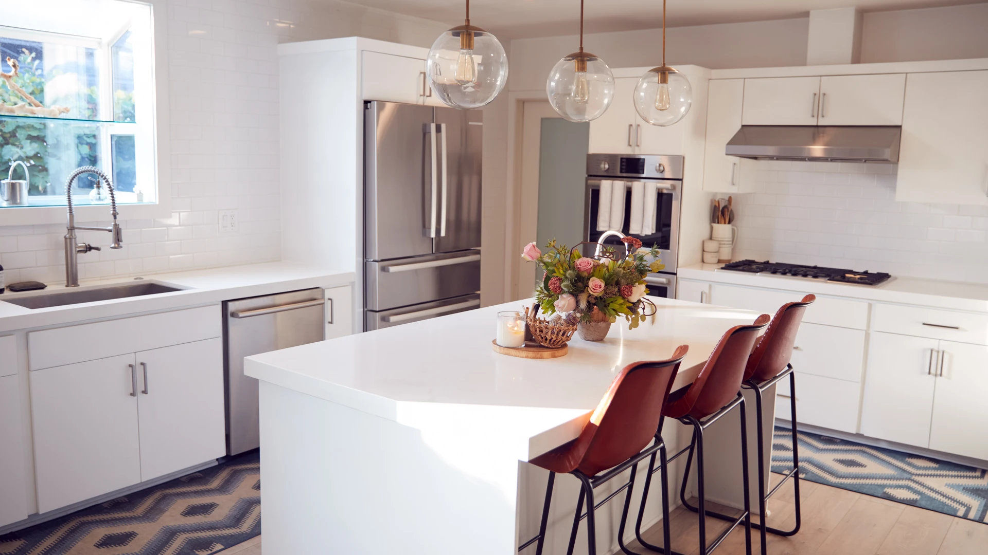 Bright white kitchen with a waterfall island, reddish-brown counter stools, globe pendants and metallic appliances.