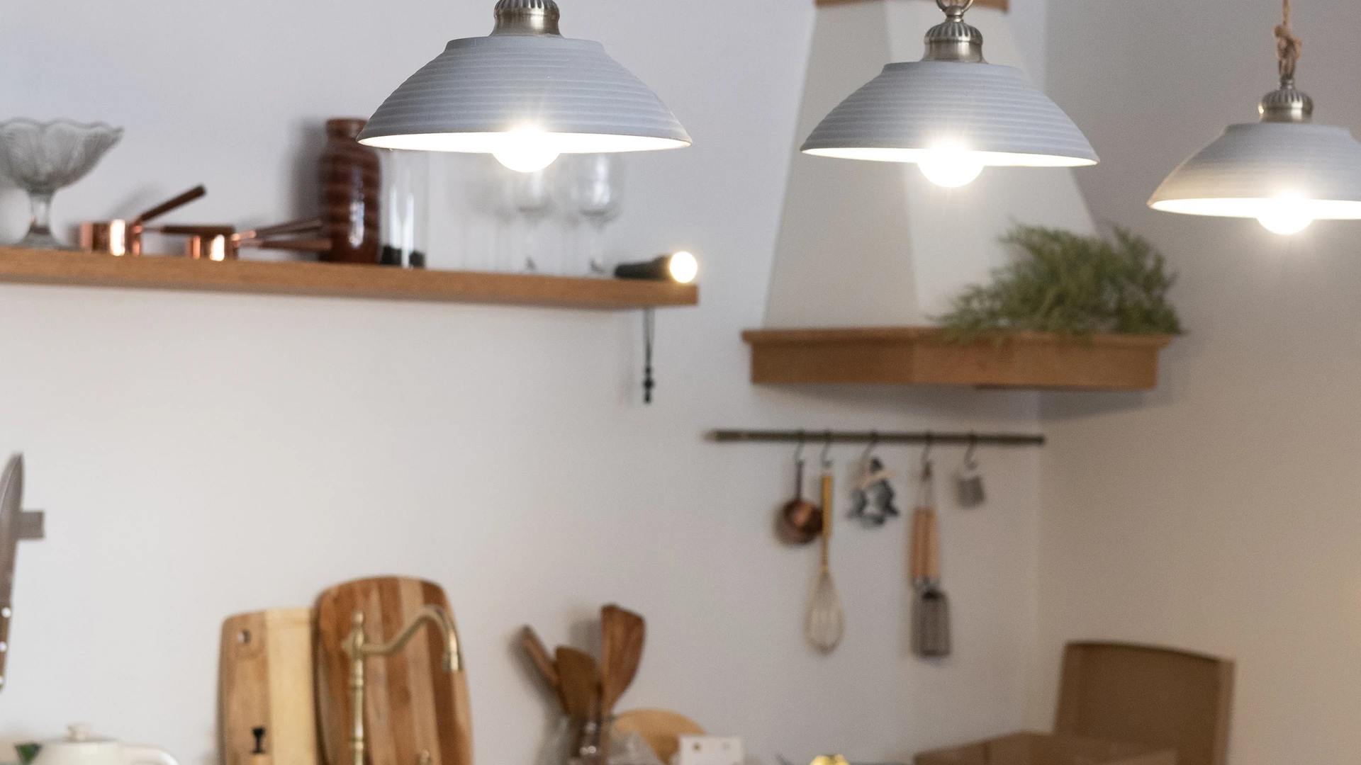 Three illuminated pendant lights with pale shades in front of open brown kitchen shelves and hanging utensils.
