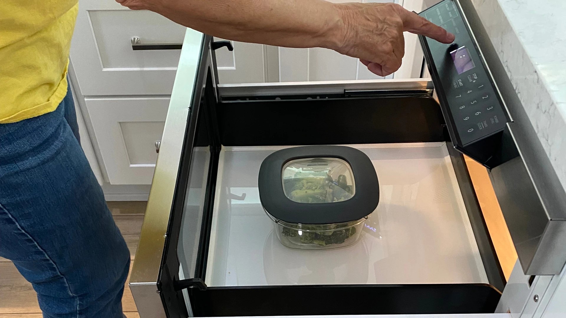 Open microwave drawer built in below the counter with a covered glass dish inside, as a person presses its control panel beside white shaker cabinets