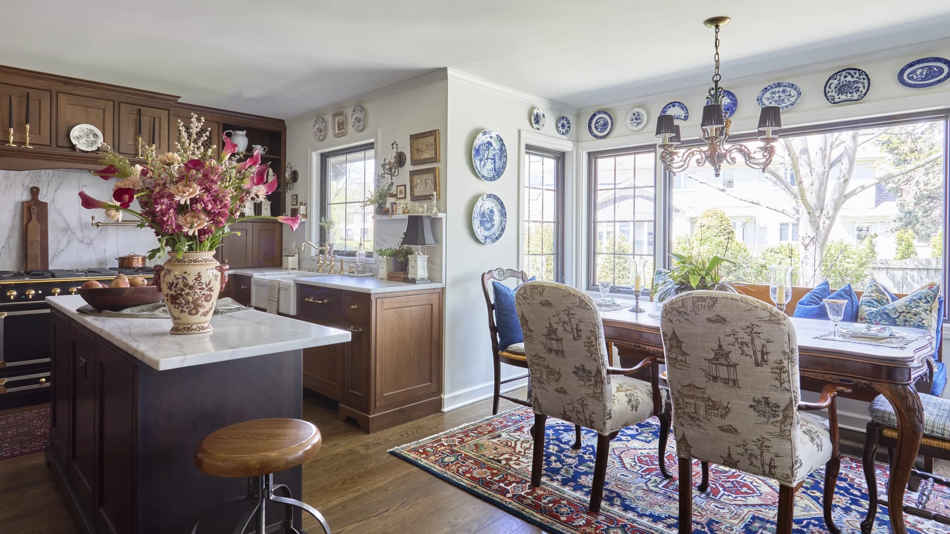 Kitchen with warm brown wood cabinets, a dark island with a white veined counter and a wooden stool, and a dining table with patterned chairs beside a bright window