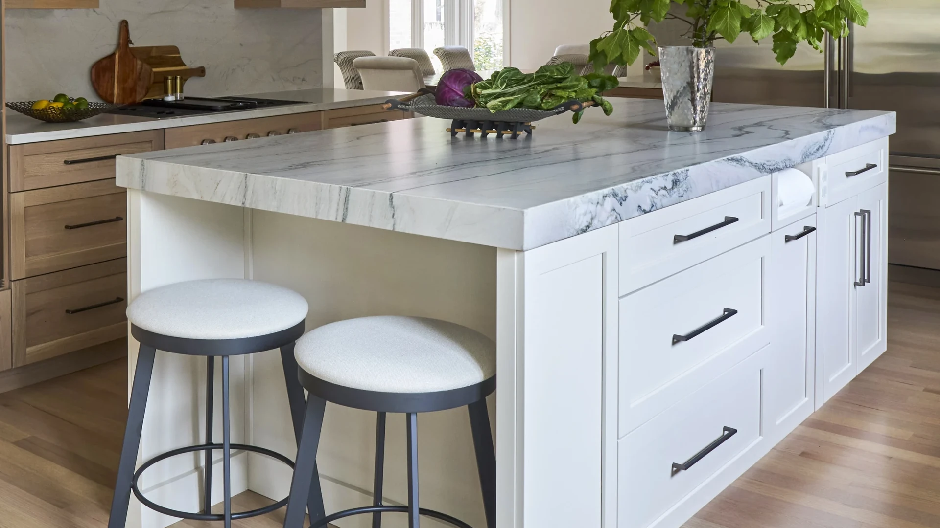 White kitchen island with a thick veined stone countertop and two round-seat stools at one end, with light wood cabinets and a cooktop beyond
