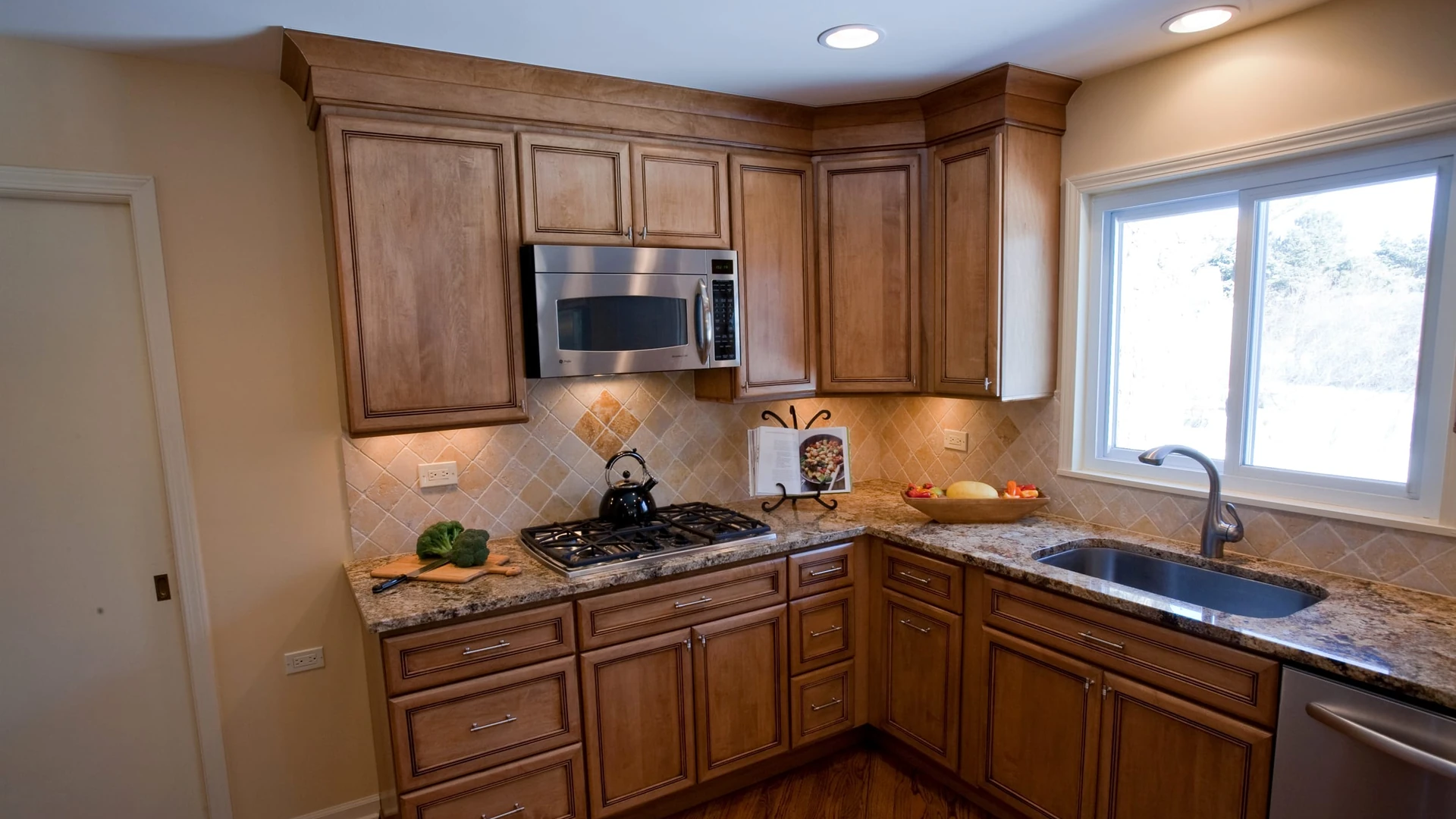 Brown wood cabinets meeting at a corner, with a gas cooktop under a microwave on one wall and a stainless steel sink under a window on the other