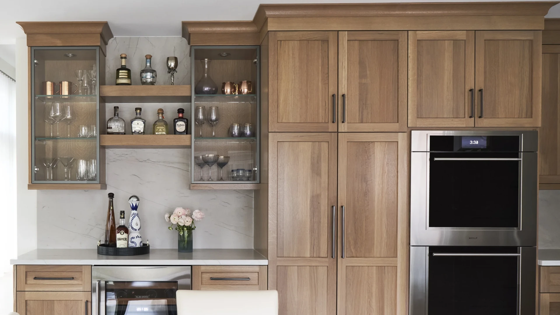 Light wood-tone cabinet wall with two glass-front wall cabinets above a beverage counter, beside tall cabinets and a stacked pair of wall ovens
