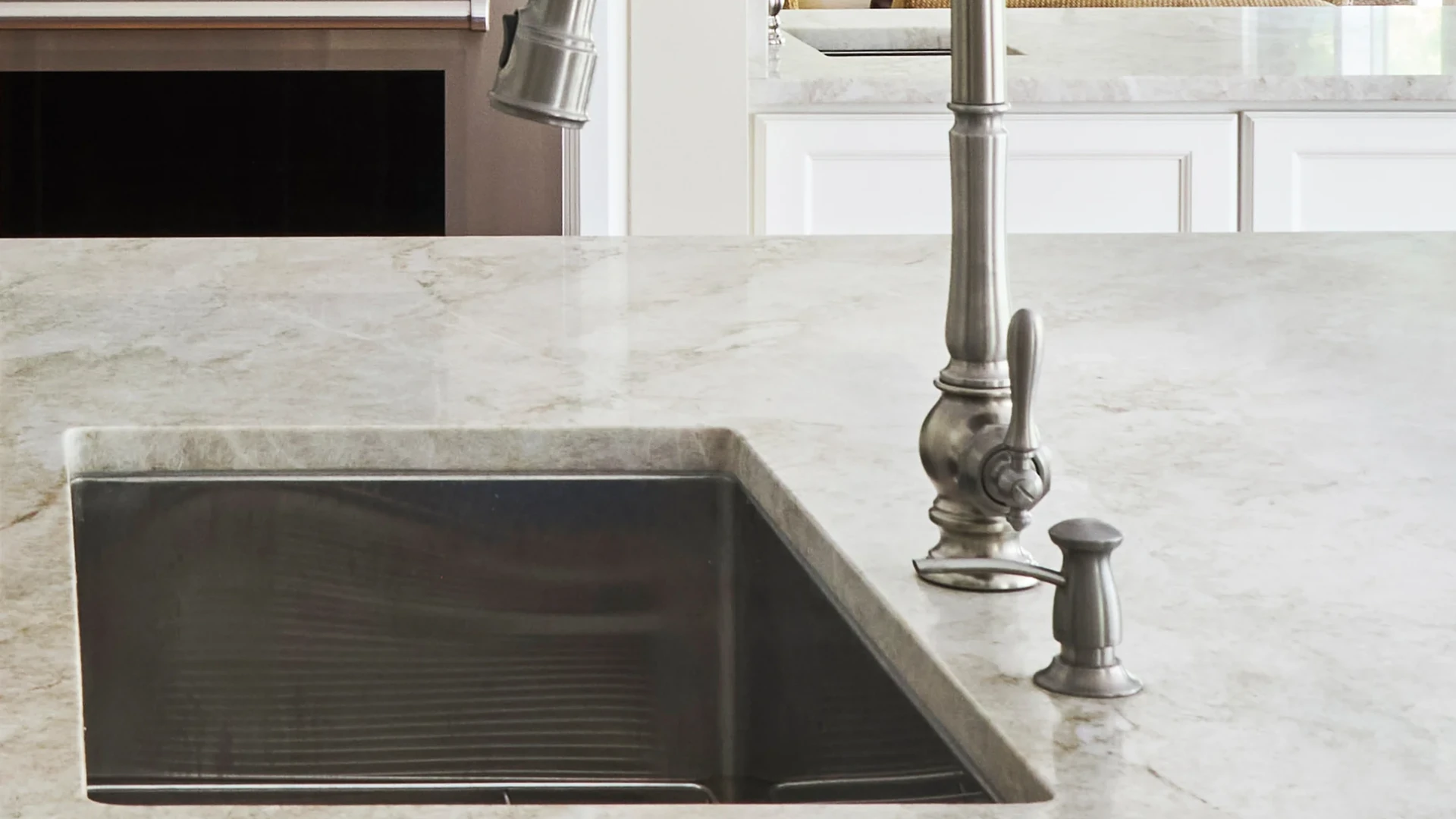 Rectangular undermount sink beside a tall faucet and soap dispenser, set in a pale veined stone countertop with white cabinets behind