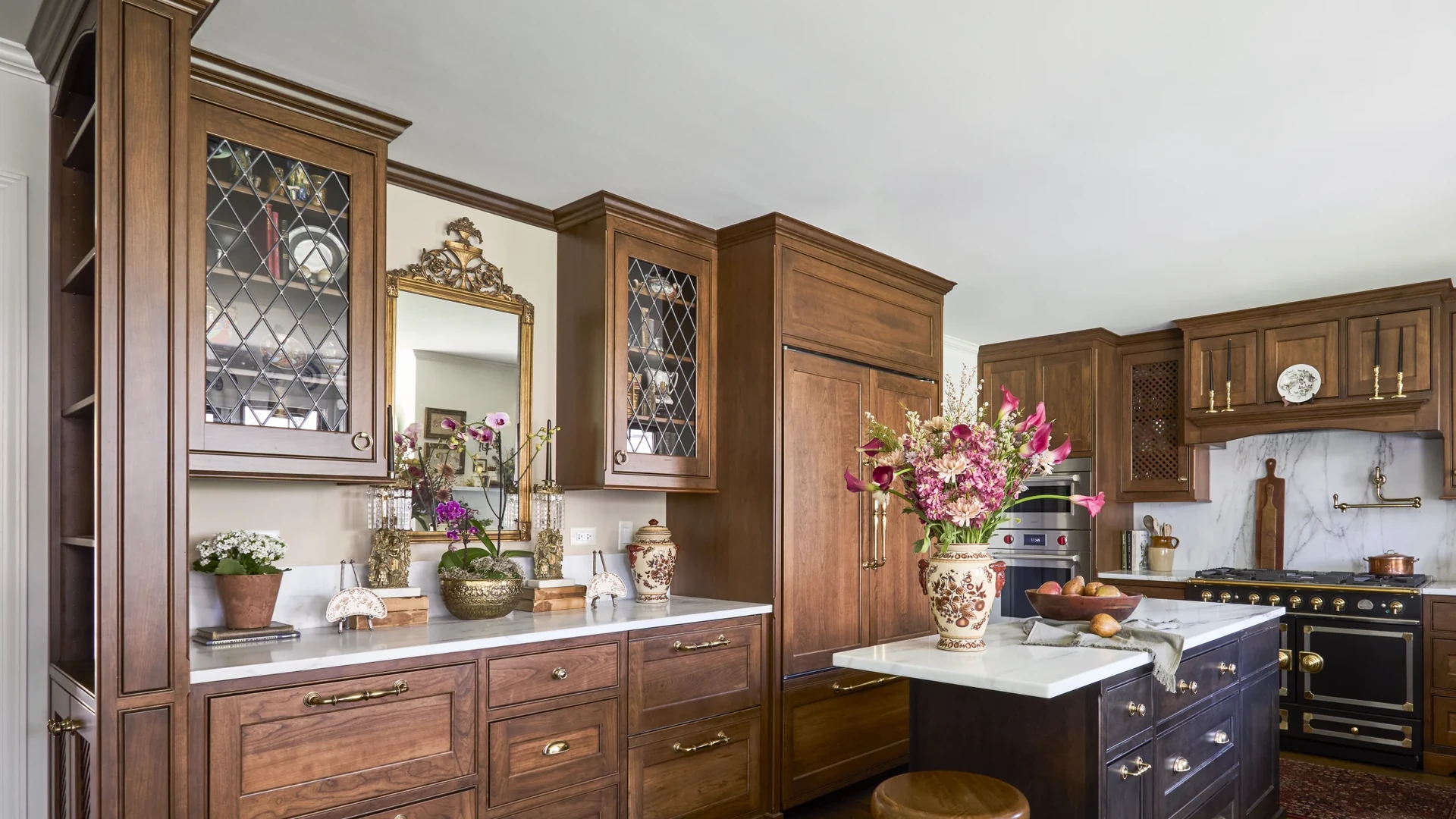 Brown wood kitchen cabinets with diamond-pattern glass upper doors and crown molding, a tall paneled cabinet and a dark island, with bare ceiling above the cabinet tops