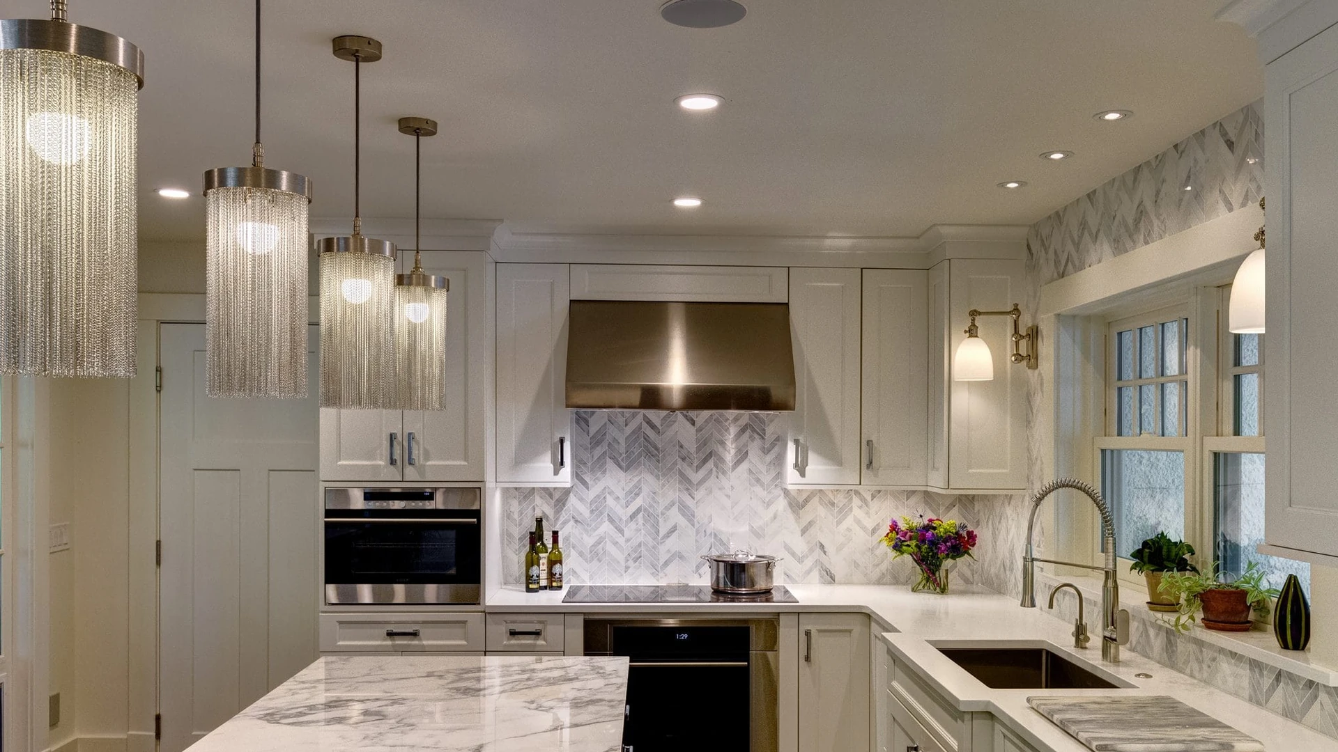 Brushed-metal hood set into a run of white upper cabinets over a black cooktop, with chevron tile behind it, pendant lights at left and a sink at right