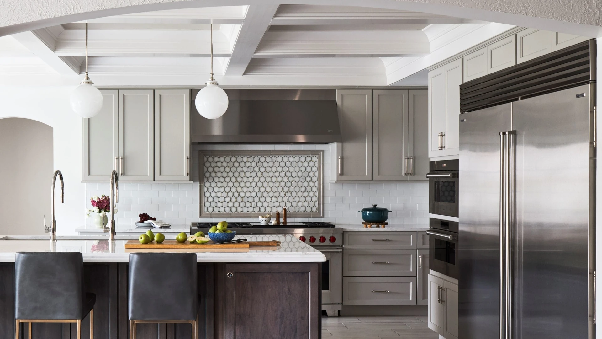 Gray wall cabinets above a white tile backsplash with a hexagon-tile panel behind a range and stainless hood, beside a dark island with two stools