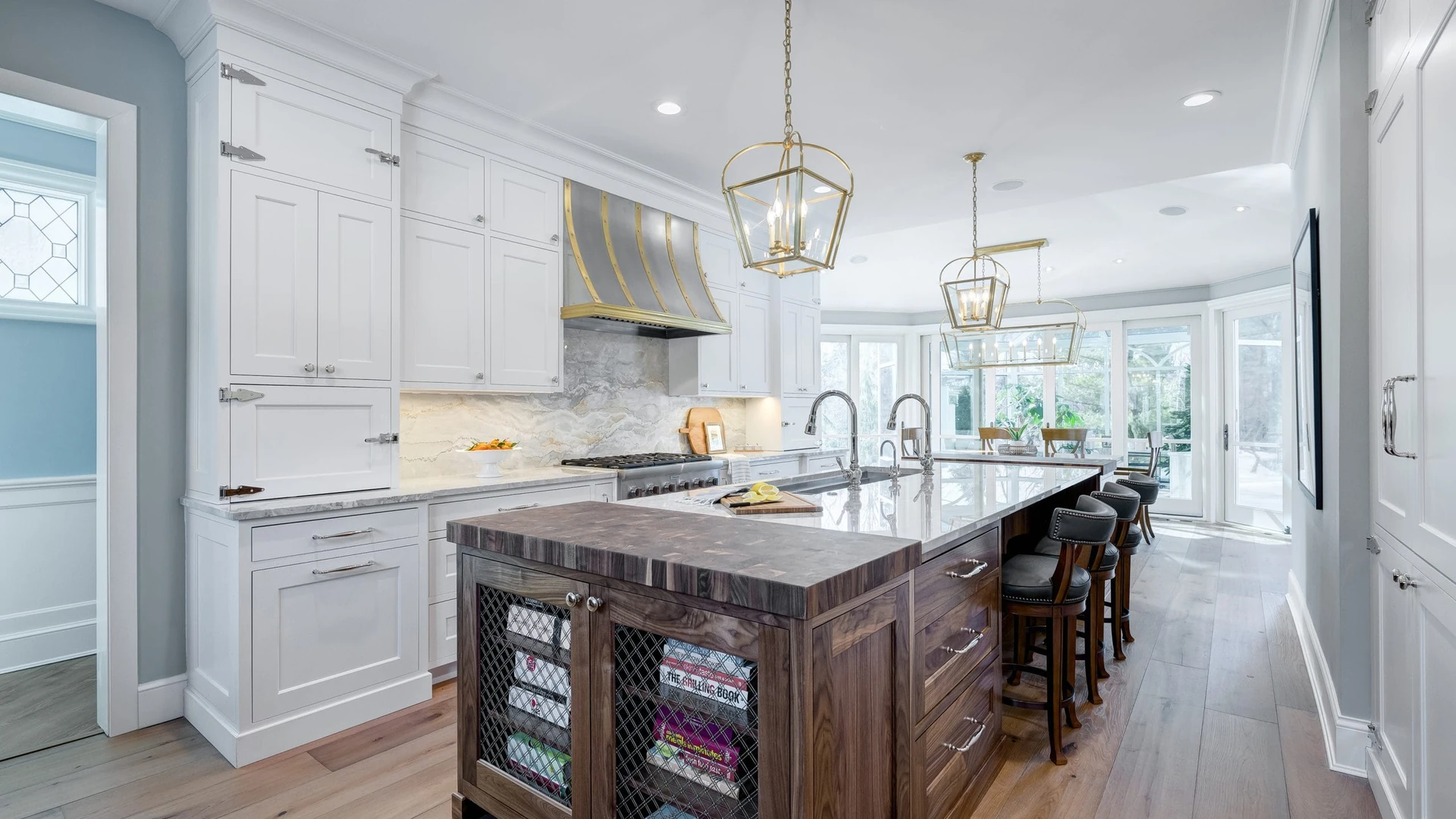 Kitchen with white painted cabinets and a long island whose dark wood cabinets have mesh-front doors and an end-grain wood top, under brass lantern pendants and a metal range hood with brass straps