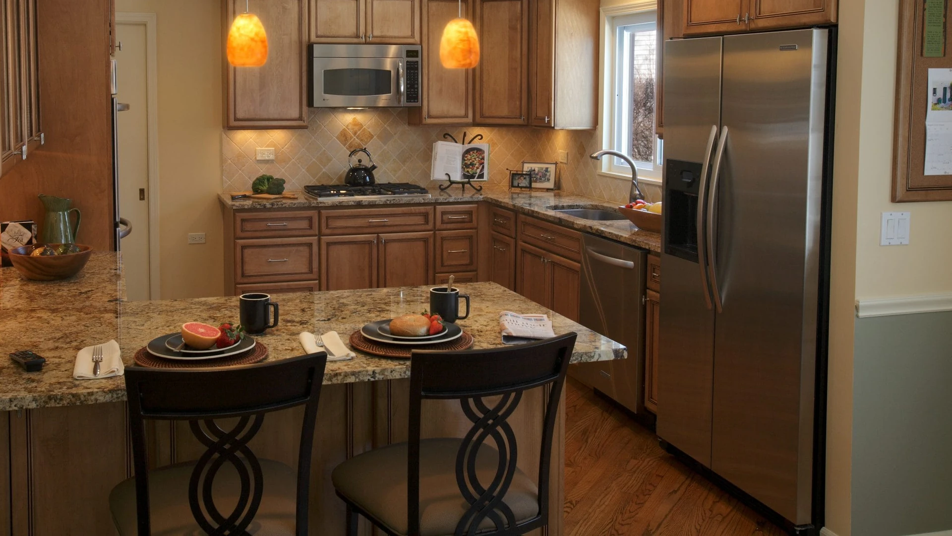 Stainless steel side-by-side refrigerator at the end of a run of brown wood cabinets, with a granite peninsula and two stools in front