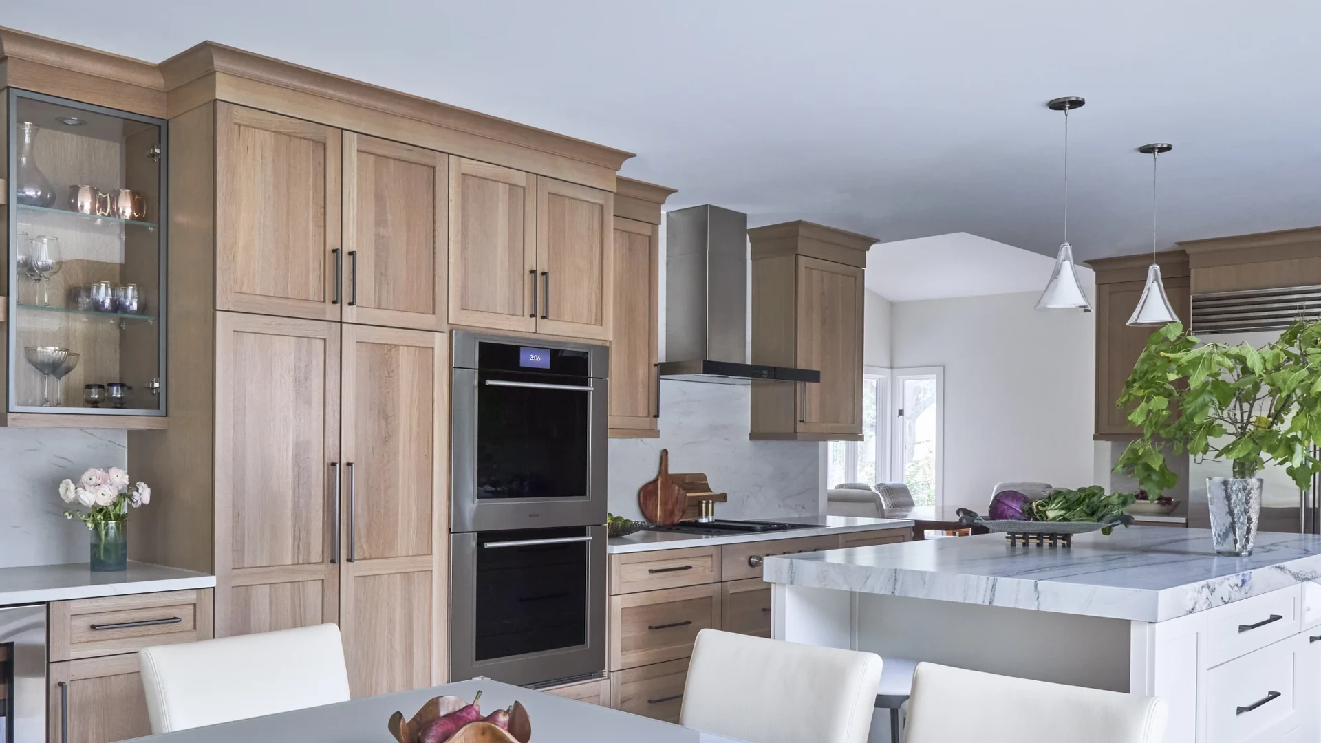 Natural-finish wood cabinets with straight vertical grain around a double wall oven and a stainless hood, with a white island and a veined stone counter at right