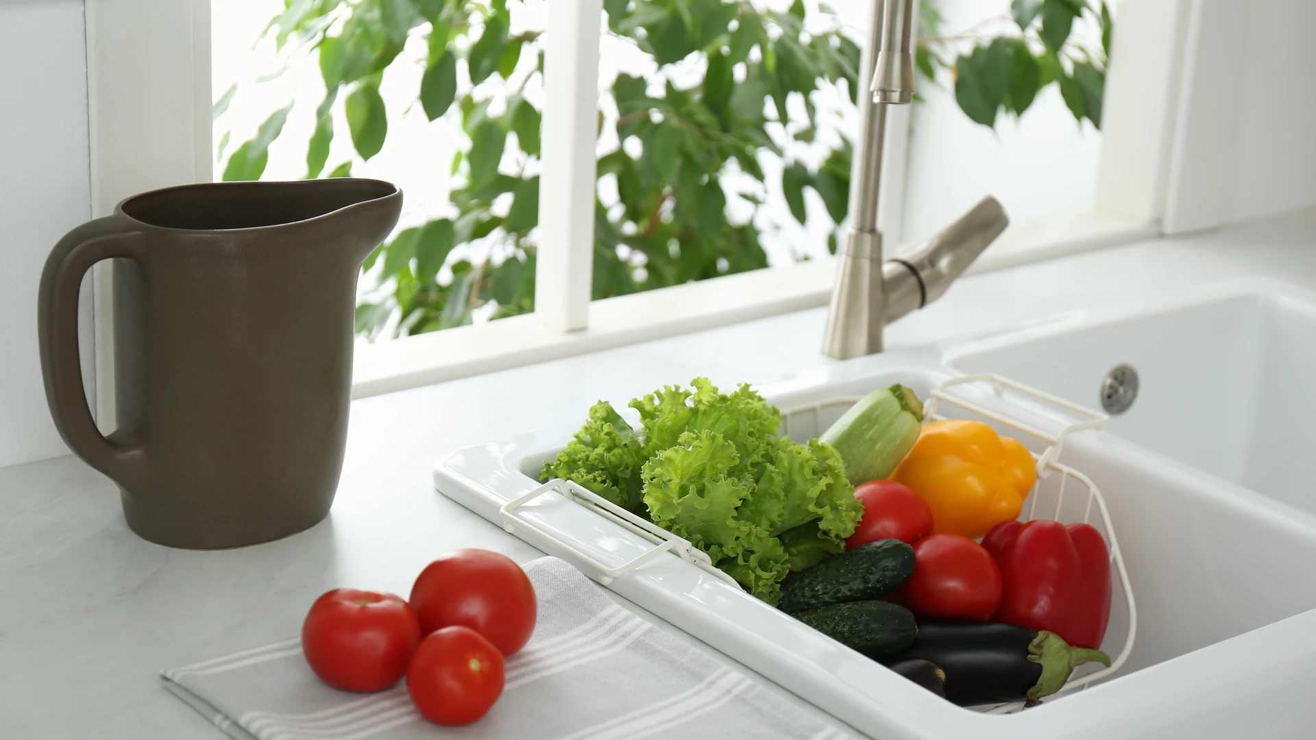A white kitchen sink with a rinse basket set across the bowl and filled with lettuce, peppers, cucumbers, tomatoes and an eggplant, beside a brown pitcher and three tomatoes on a striped towel